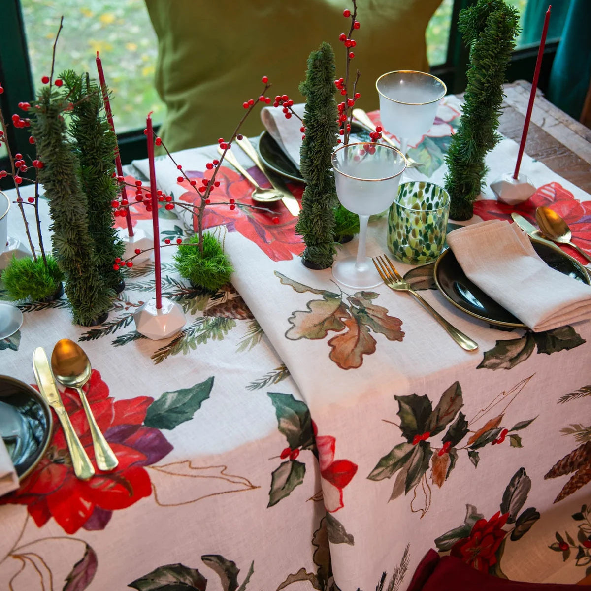 A festive holiday table setting featuring a white tablecloth printed with red poinsettias, green holly leaves, and gold outlines. The table is decorated with miniature green Christmas trees, red berry garlands, gold flatware, white plates, and folded white napkins.
