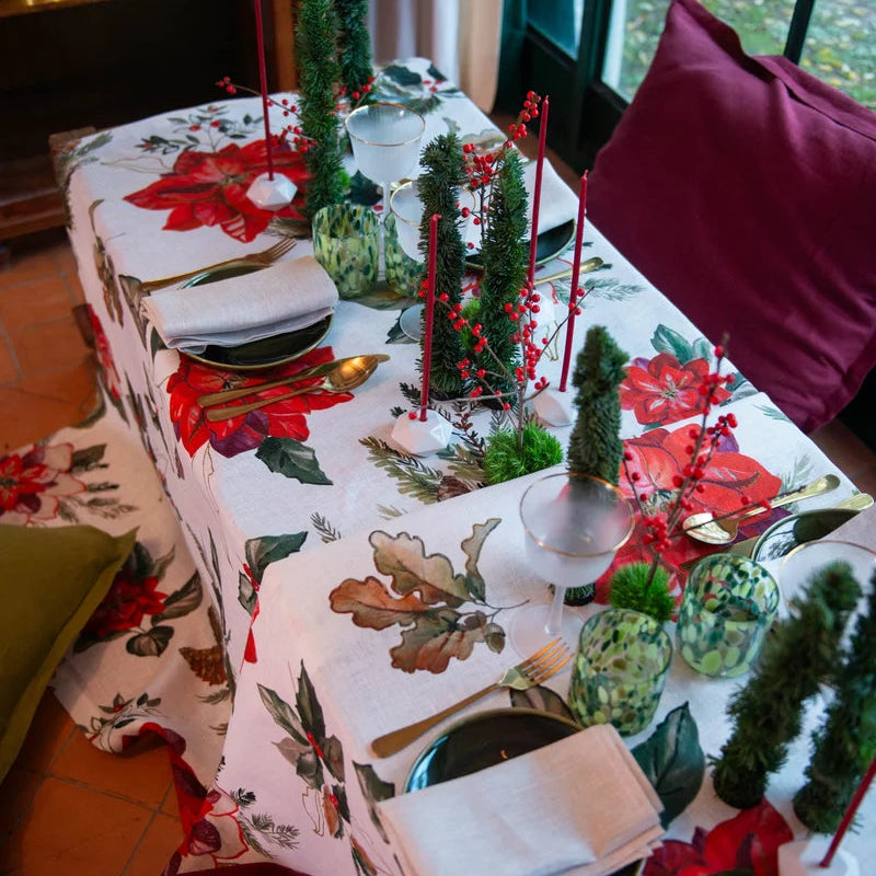 A festive holiday table setting featuring a white tablecloth printed with large red poinsettias, green oak leaves, pinecones, and pine trees. The table is set with black plates, white napkins, gold cutlery, and decorative green glass vases containing miniature pine trees and red candlesticks.